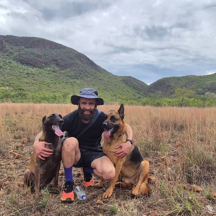 Guy Carosin kneeling between his Belgian Malinois and German Shepherd in the bushveld